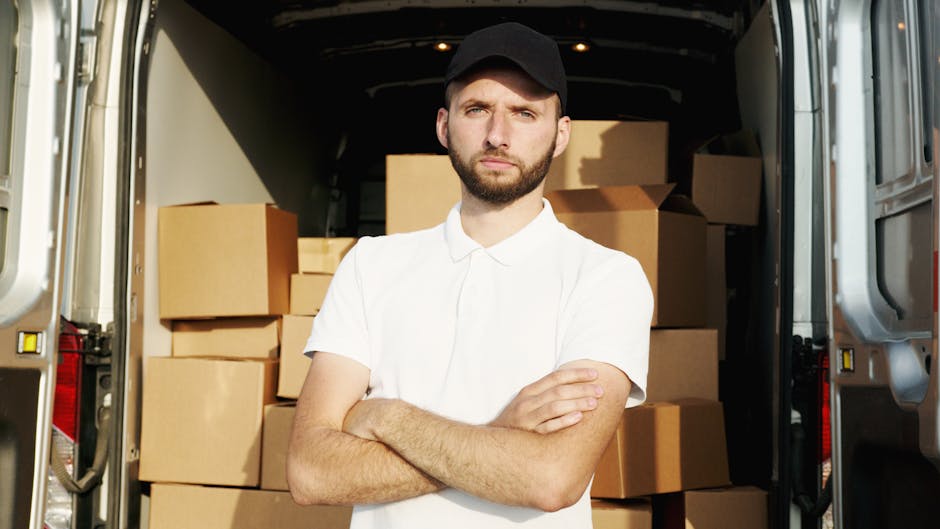 A young man with a serious expression, wearing a plain white polo shirt and a black cap, stands inside the open rear of a moving van surrounded by neatly stacked cardboard boxes of various sizes, some sealed with packing tape, in an indoor setting with dim lighting. The boxes are arranged to maximize space efficiency for home relocation, with the van's interior visible behind him showing additional packing supplies and equipment used in furniture transport and packing and moving processes. The man appears to be overseeing the loading process, which involves careful handling and spacing of the boxes to ensure safe transportation during the house move, as part of professional removals services offered by Man with Van Brent. The scene emphasizes logistics, packing materials such as cardboard, and the strategic organization required in house removals and furniture transport.