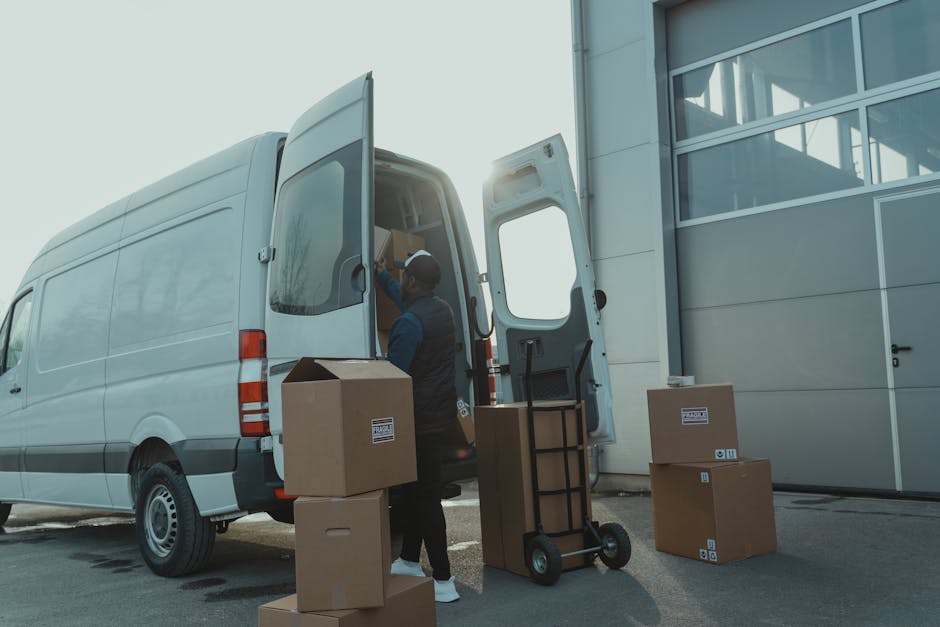 A professional mover from Man with Van Brent is loading several cardboard boxes and a small moving trolley into the back of a large white van outside a modern commercial building. The boxes, made of brown corrugated cardboard, vary in size and are stacked on the pavement near the vehicle. The mover, dressed in dark work clothes and wearing a baseball cap, is placing items inside the van, which has its rear doors open, revealing a partially visible interior used for transport. A few more packed boxes are positioned on the ground nearby, ready for home relocation or furniture transport. The setting is outdoors, with natural daylight illuminating the scene, emphasizing the loading process that is part of effective packing and moving logistics, typical of house removal services offered by Man with Van Brent. The scene highlights the careful handling and organization involved in completing a house move efficiently.