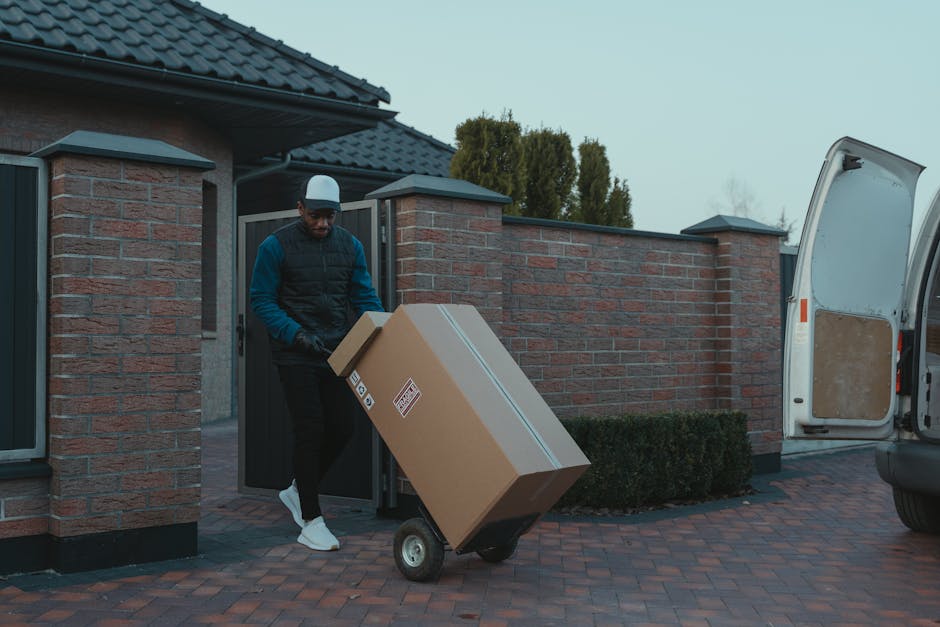 A young man with a serious expression, wearing a plain white polo shirt and a black cap, stands inside the open rear of a moving van surrounded by neatly stacked cardboard boxes of various sizes, some sealed with packing tape, in an indoor setting with dim lighting. The boxes are arranged to maximize space efficiency for home relocation, with the van's interior visible behind him showing additional packing supplies and equipment used in furniture transport and packing and moving processes. The man appears to be overseeing the loading process, which involves careful handling and spacing of the boxes to ensure safe transportation during the house move, as part of professional removals services offered by Man with Van Brent. The scene emphasizes logistics, packing materials such as cardboard, and the strategic organization required in house removals and furniture transport.