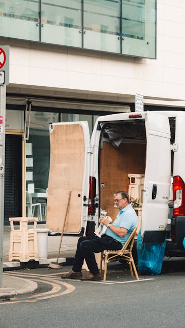 A man with glasses, wearing a light blue shirt and dark trousers, is seated on a wooden chair next to a white delivery van parked on a urban street. The van's rear doors are open, revealing a partially loaded interior with cardboard boxes, wooden furniture, and packing materials. The man appears to be resting or reviewing details, holding a piece of paper or a checklist. Nearby, small wooden stools and protective padding are visible on the pavement, while the street background includes a modern building with glass windows and a no-parking traffic sign. The scene illustrates a home relocation process involving furniture transport and packing, with the man part of a professional removals service such as Man with Van Brent, positioning the vehicle for efficient loading or unloading during a house move or flat removal in Kilburn.