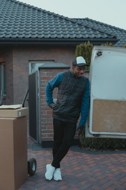 A man stands outdoors next to an open white moving van parked on a paved driveway in front of a residential house with grey tiled roofing and brick walls. He is wearing a black padded vest over a blue long-sleeved shirt, black trousers, white shoes, and has a grey cap. The man is leaning against the van with one hand on his hip and appears to be observing the loading process. Nearby, there are large cardboard boxes, one on a small trolley, which are likely packed for a home relocation. The scene depicts a typical moving day environment, with the man appearing to be part of a professional removals service such as Man with Van Brent, engaged in packing, loading, or transporting furniture and household items using protective blankets or wrapping materials visible on some objects. The outdoor setting is well-lit with natural daylight, highlighting the preparation steps involved in furniture transport and packing during house removals.