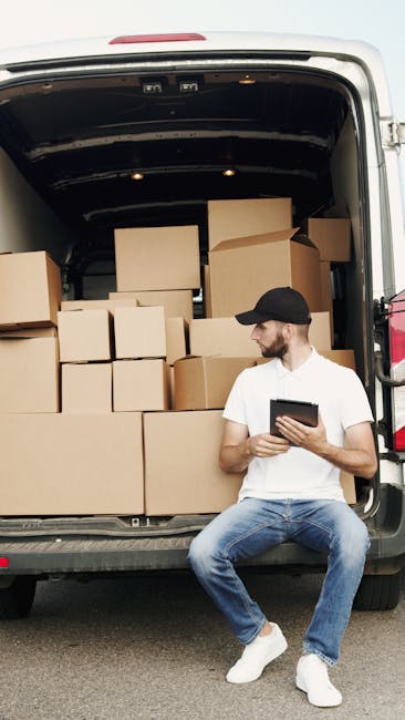 A man with glasses, wearing a light blue shirt and dark trousers, is seated on a wooden chair next to a white delivery van parked on a urban street. The van's rear doors are open, revealing a partially loaded interior with cardboard boxes, wooden furniture, and packing materials. The man appears to be resting or reviewing details, holding a piece of paper or a checklist. Nearby, small wooden stools and protective padding are visible on the pavement, while the street background includes a modern building with glass windows and a no-parking traffic sign. The scene illustrates a home relocation process involving furniture transport and packing, with the man part of a professional removals service such as Man with Van Brent, positioning the vehicle for efficient loading or unloading during a house move or flat removal in Kilburn.