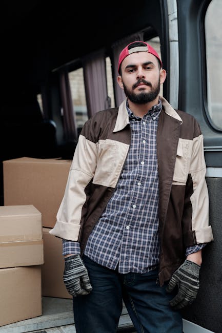 A man dressed in casual clothing, including a white t-shirt, blue jeans, white sneakers, and a black cap, is sitting on the ground next to an open delivery van during a home relocation process. The va